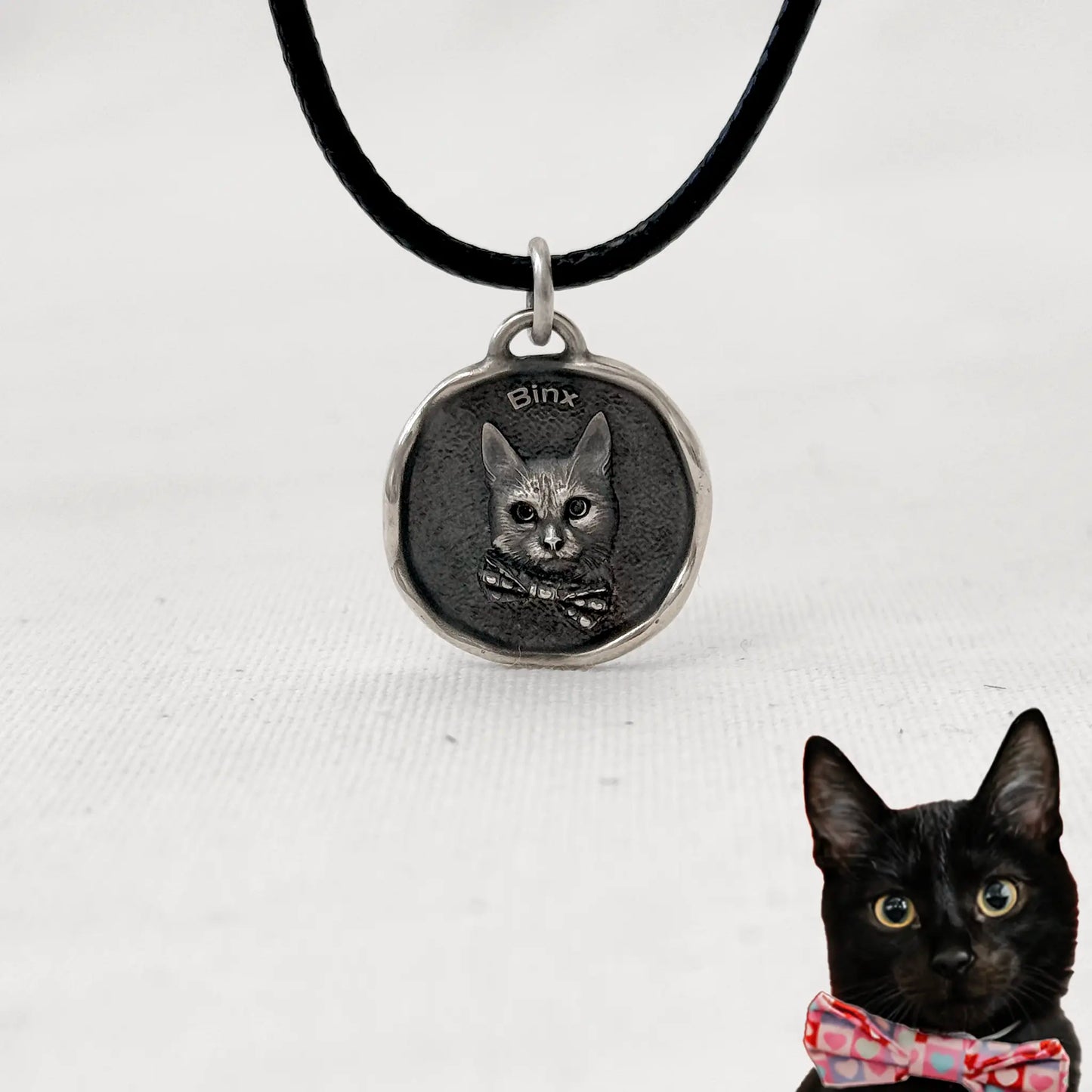Custom silver cat pendant with engraved portrait of Binx, wearing a bow tie, on a black cord necklace.