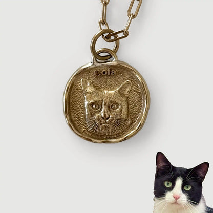 Gold pendant with a cat's face engraving and a real cat below on a white background