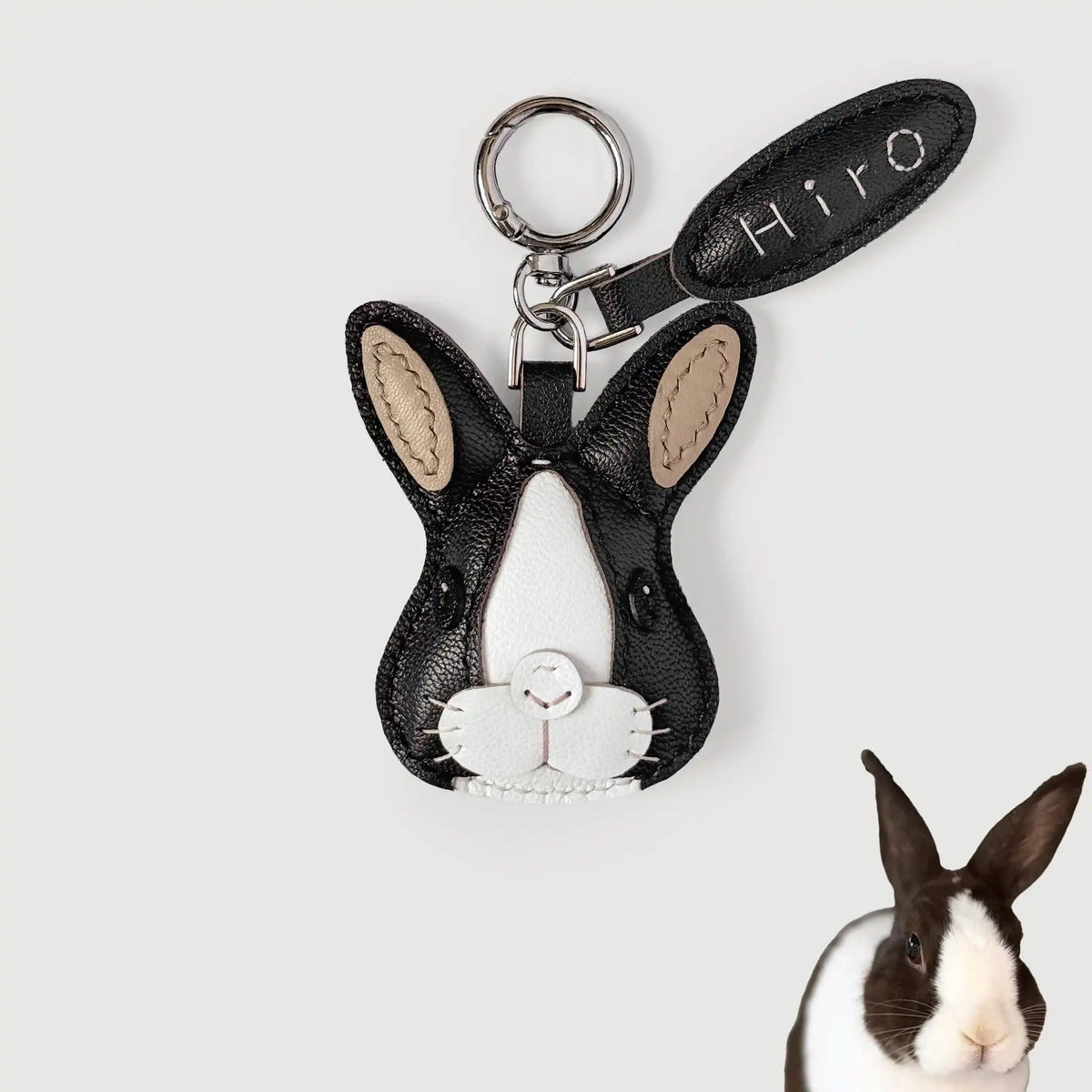 A black and white rabbit-shaped keychain charm with beige ear inlays and a silver key ring, shown on a light gray background with a small rabbit photo in the lower right corner. The leather tag reads 'Hiro'.
