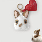 A white faux-leather rabbit plush keychain with tan ears, black eyes, and a red heart-shaped tag embroidered with the name 'Luca'. A small real rabbit is partially visible in the bottom-right corner against a light gray background.