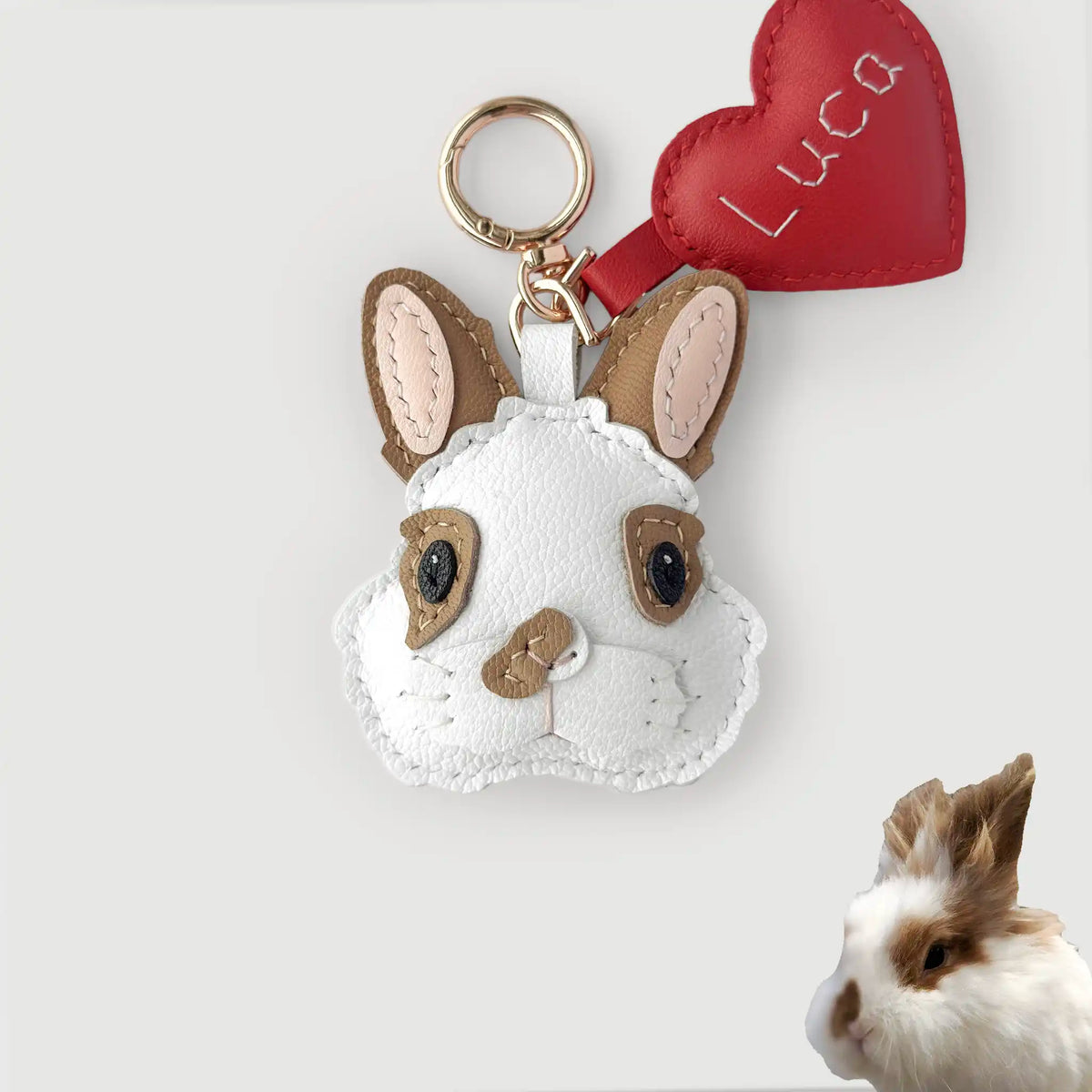 A white faux-leather rabbit plush keychain with tan ears, black eyes, and a red heart-shaped tag embroidered with the name 'Luca'. A small real rabbit is partially visible in the bottom-right corner against a light gray background.