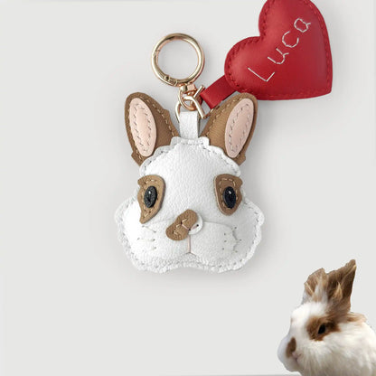 A white faux-leather rabbit plush keychain with tan ears, black eyes, and a red heart-shaped tag embroidered with the name 'Luca'. A small real rabbit is partially visible in the bottom-right corner against a light gray background.