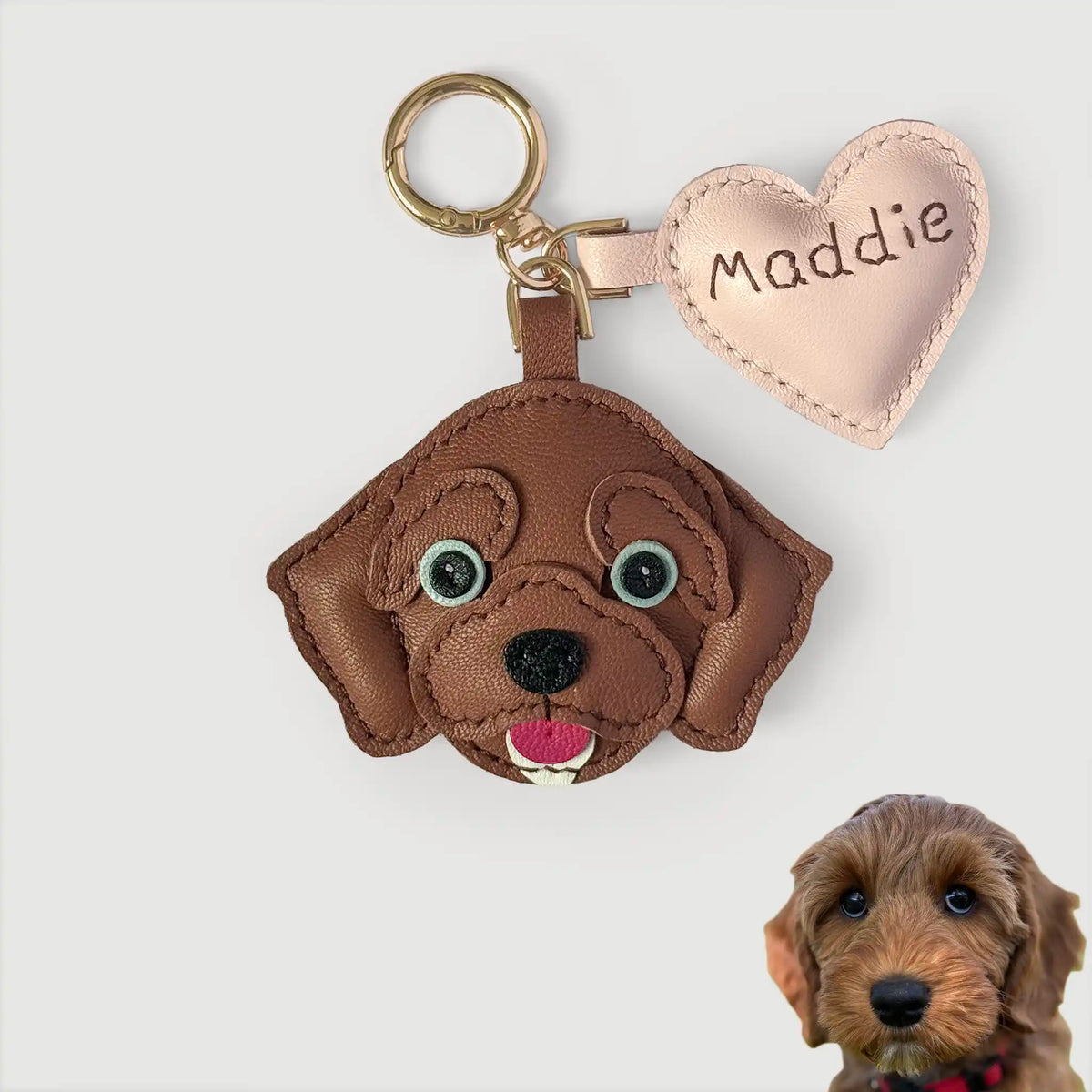 A brown leather dog-shaped keychain with a gold split ring and a light pink heart tag engraved with the name 'Maddie'. A small photo of a real dog is shown in the bottom right corner.