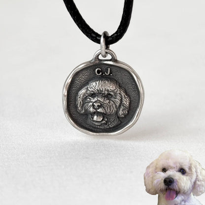 Silver pendant with a dog's face and initials, attached to a black cord, with a white dog in the background.