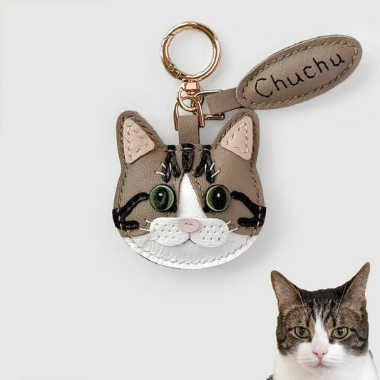 A handmade cat-face keychain charm made from brown and white leather, featuring embroidered black stripes, green bead eyes, and a gold key ring. A matching leather tag is attached with the name 'Chuchu' stitched on it.