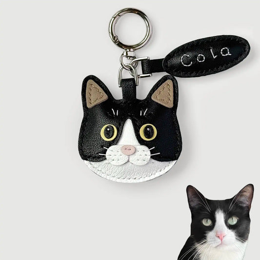 A black and white cat face keychain charm with yellow eyes, a silver key ring, and a black leather tag reading 'Cola' on a light gray background. A small photo of a real black-and-white cat is shown in the bottom right corner.