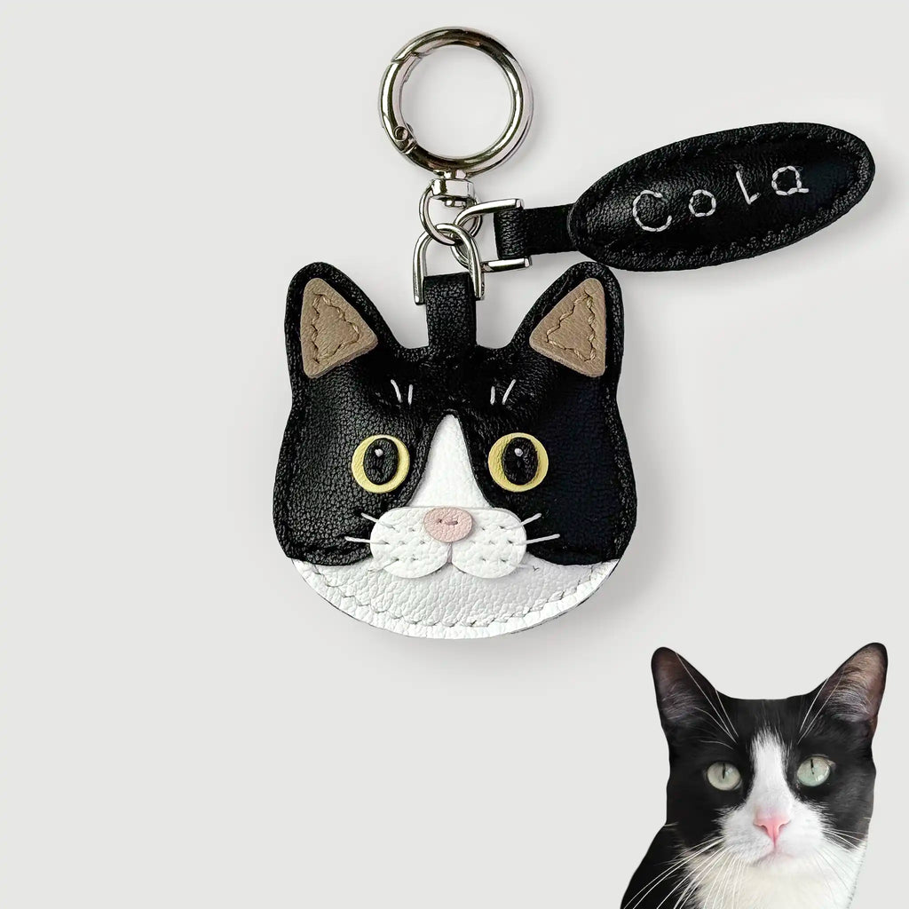 A black and white cat face keychain charm with yellow eyes, a silver key ring, and a black leather tag reading 'Cola' on a light gray background. A small photo of a real black-and-white cat is shown in the bottom right corner.