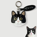 A black and white cat face keychain charm with yellow eyes, a silver key ring, and a black leather tag reading 'Cola' on a light gray background. A small photo of a real black-and-white cat is shown in the bottom right corner.