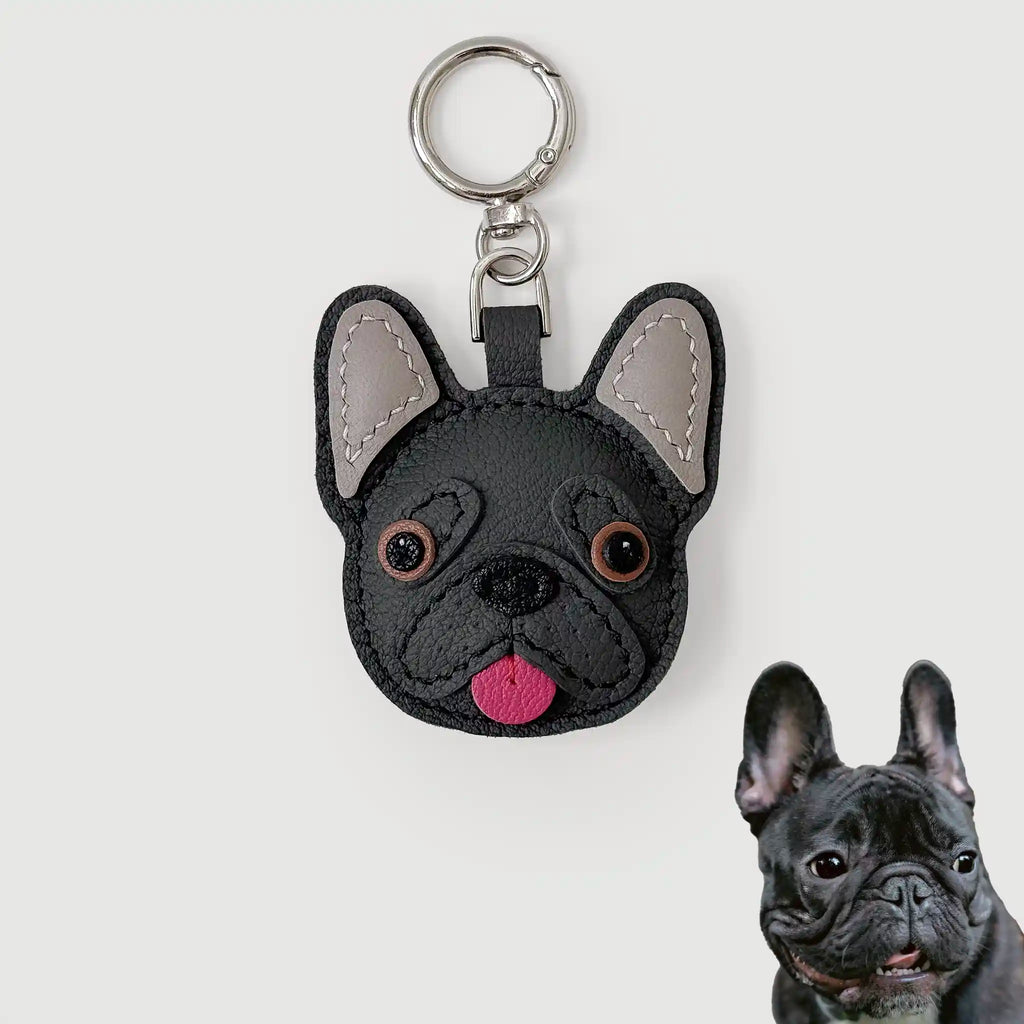 A black faux-leather French Bulldog keychain with gray inner ears, brown eyes, and a pink tongue, shown on a light gray background with a small inset photo of a real French Bulldog in the bottom right corner.