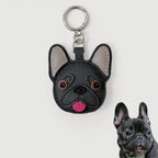 A black faux-leather French Bulldog keychain with gray inner ears, brown eyes, and a pink tongue, shown on a light gray background with a small inset photo of a real French Bulldog in the bottom right corner.