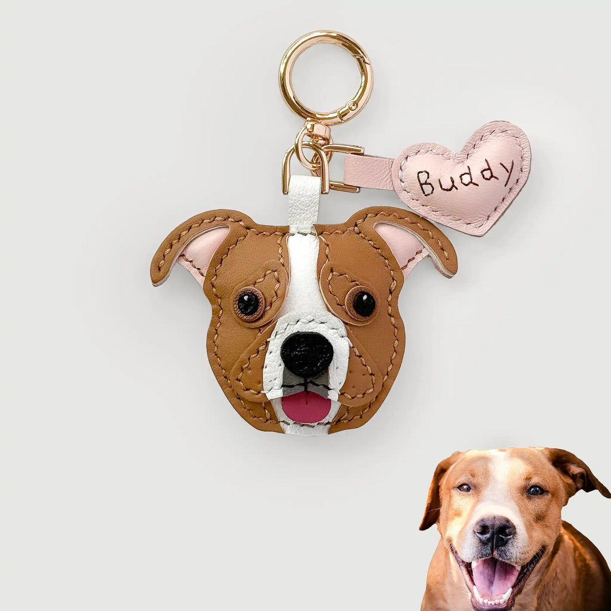 Custom leather pet keychain featuring a dog head cutout with brown and white leather, a gold key ring, and a pink heart-shaped tag labeled 'Buddy'. A real dog photo is shown in the bottom right corner against a light gray background.