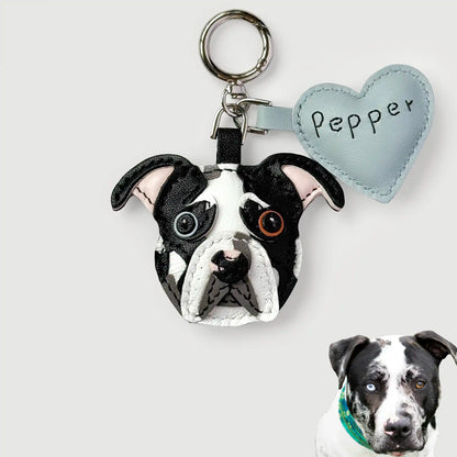 A custom dog keychain featuring a realistic black and white bulldog face with embroidered details, attached to a silver key ring and a light blue heart-shaped tag labeled 'Pepper'. A small inset photo of a real black and white dog wearing a green bandana is shown in the bottom right corner.