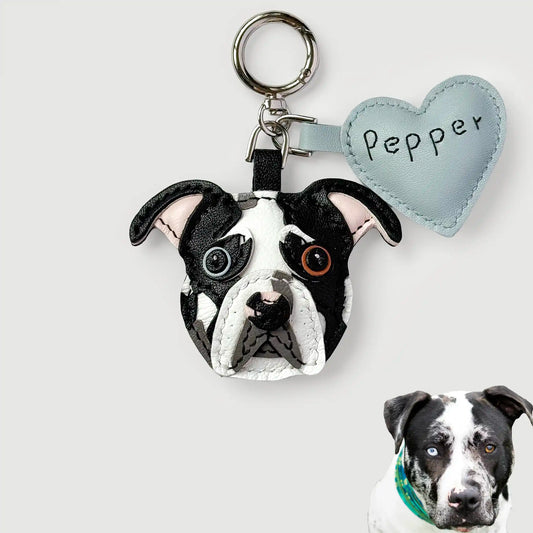 A custom dog keychain featuring a realistic black and white bulldog face with embroidered details, attached to a silver key ring and a light blue heart-shaped tag labeled 'Pepper'. A small inset photo of a real black and white dog wearing a green bandana is shown in the bottom right corner.